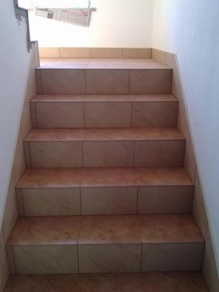 Wykończone schody wewnętrzne z jasnymi, kwadratowymi płytkami ceramicznymi. Widok z dołu, na ścianie gazeta zabezpieczona folią malarską.