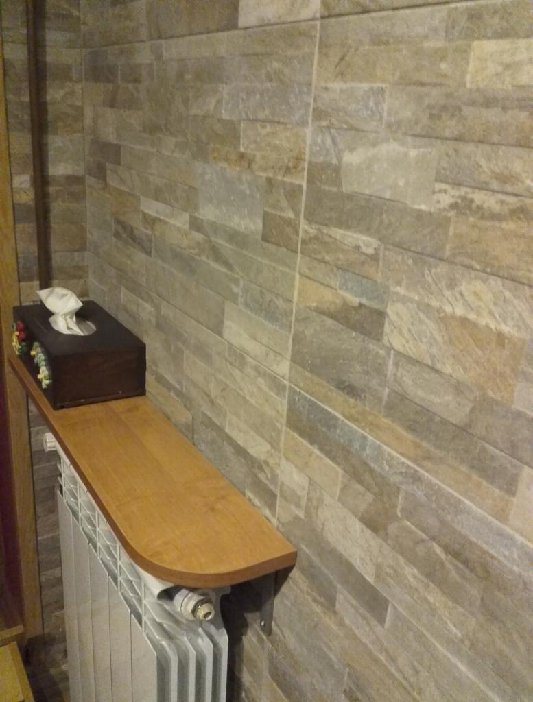 Wykończona łazienka z kamienną okładziną ściany, drewnianą półką nad grzejnikiem i ozdobnym pudełkiem na chusteczki. Detal wykończenia wnętrza.