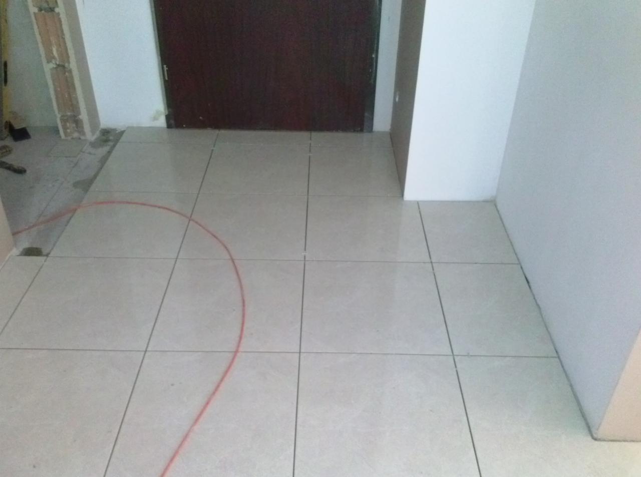 Jasne, kwadratowe płytki podłogowe po ułożeniu w korytarzu, widoczne fragmenty ściany i drzwi. Czerwony kabel na płytkach. Częściowo wykończone pomieszczenie.