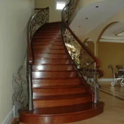 Zakrzywione schody drewniane z ozdobną, metalową balustradą z motywem roślinnym, widziane z dołu, z parkietem w tle. Ciepłe oświetlenie.