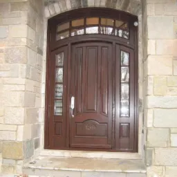 Eleganckie, ciemnobrązowe drzwi zewnętrzne z łukowym nadświetlem i przeszkleniami bocznymi, wkomponowane w kamienną fasadę z oświetleniem zewnętrznym.