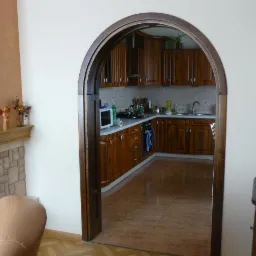 Kuchnia z drewnianymi meblami widziana przez łukowe przejście. W tle widoczny fragment kominka z dekoracjami. Ciepłe oświetlenie wnętrza.