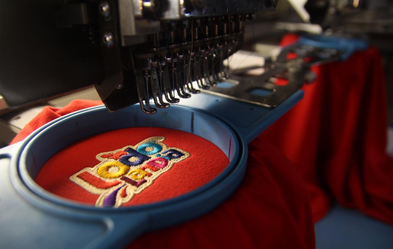 Haftowanie logo na czerwonej koszulce za pomocą profesjonalnej maszyny hafciarskiej. Precyzyjne detale i żywe kolory. Gotowy produkt.