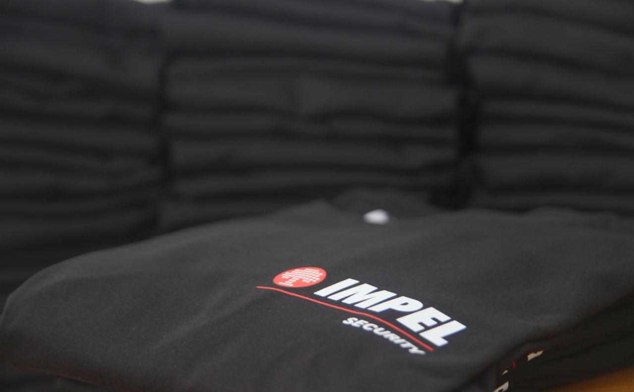 Stos czarnych koszulek z nadrukowanym logo 'IMPEL Security' w kolorze białym i czerwonym. Ułożone w stos, lekko rozmyte tło.