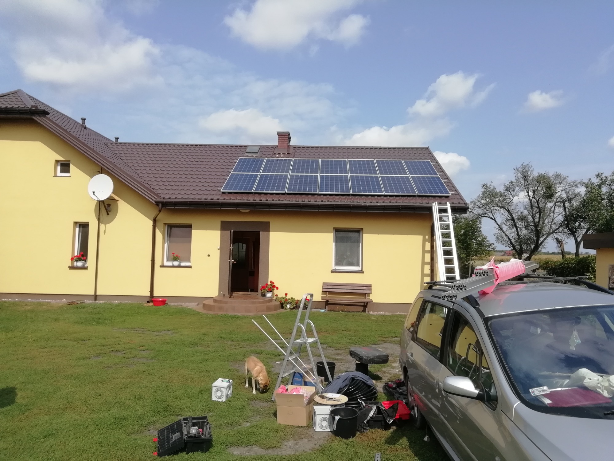 Żółty dom jednorodzinny z brązowym dachem, na którym zamontowane są panele słoneczne. Na dachu oparta drabina. Na trawniku przed domem leżą narzędzia i materiały montażowe, obok stoi srebrny...