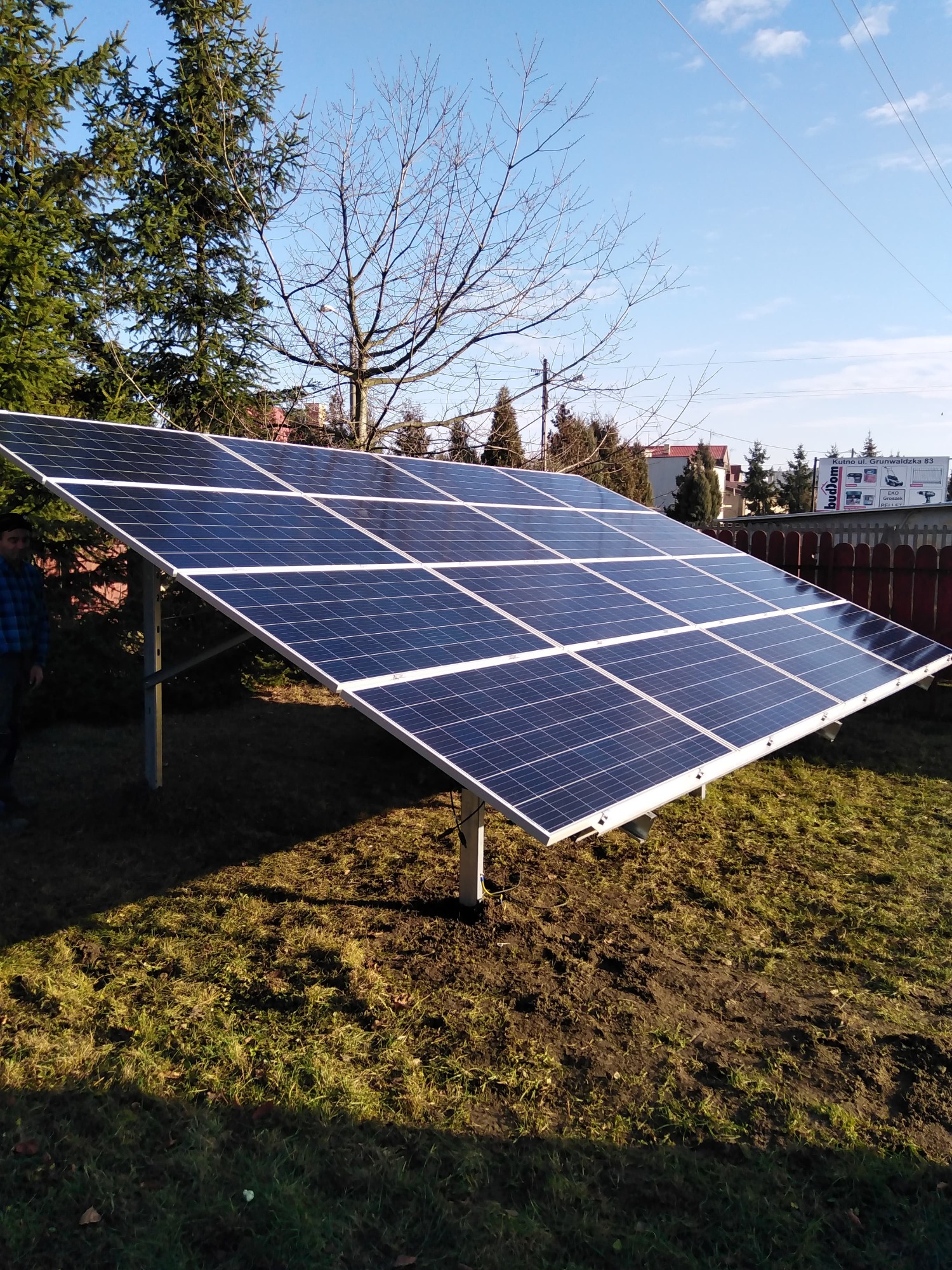 Panel słoneczny na metalowej konstrukcji ustawiony na trawniku przed domem, widoczne drzewa i niebieskie niebo w tle.