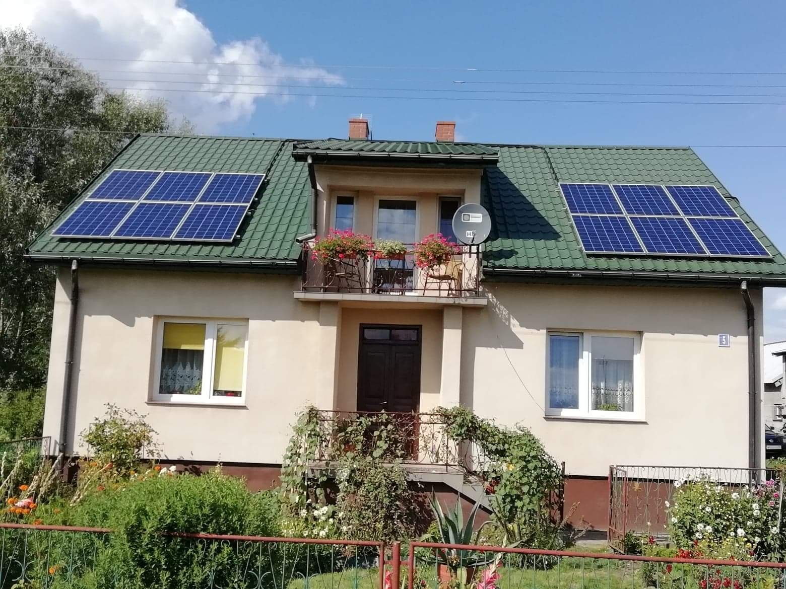 Dwupiętrowy dom z zielonym dachem i panelami słonecznymi, balkon z kwiatami, antena satelitarna.