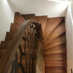 Drewniane schody spiralne z ażurową balustradą, widok z dołu, oświetlenie górne podkreśla strukturę drewna.