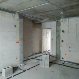 Wnętrze w stanie surowym z widocznymi przewodami elektrycznymi ułożonymi na podłodze i pod sufitem, przygotowane do instalacji gniazdek i oświetlenia.