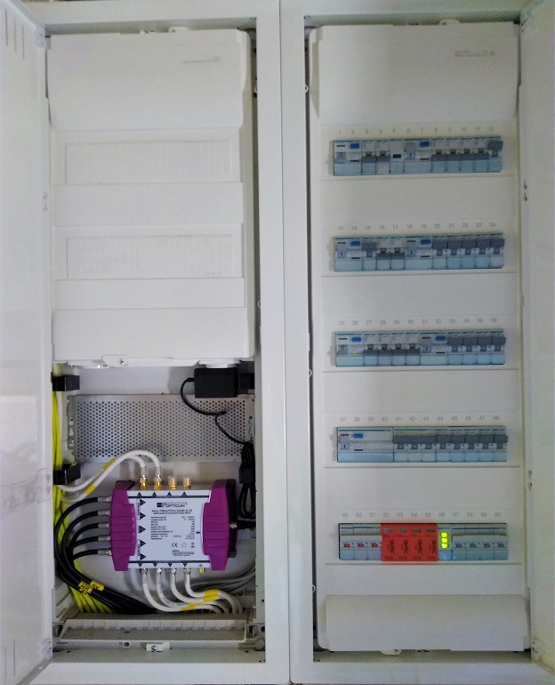 Otwarta szafa rozdzielcza z bezpiecznikami i modułem OPTICUM, widoczne okablowanie i elementy zabezpieczeń.