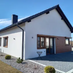 Nowoczesny dom jednorodzinny z białymi ścianami, brązowymi ramami okiennymi i zewnętrznymi roletami w kolorze drewna, widok z ogrodu na tle błękitnego nieba.