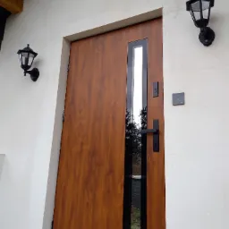 Zewnętrzne brązowe drzwi wejściowe z pionowym przeszkleniem, czarną klamką i dwoma czarnymi lampami po bokach na białej ścianie budynku.