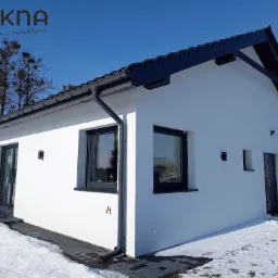 Nowoczesny, parterowy dom z białymi ścianami i czarną stolarką okienną, widok z zewnątrz w zimowej scenerii.