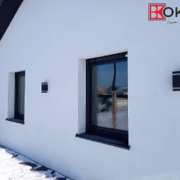 Biała elewacja domu z czarnymi oknami aluminiowymi, lampami i zbiornikiem na deszczówkę w zimowej scenerii, widoczne logo firmy 'Okna Nowa Perspektywa'.