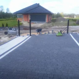 Fragment podjazdu z ciemnej kostki brukowej z białymi pasami, w tle budynek w trakcie budowy, zagęszczarka wibracyjna i materiały budowlane.