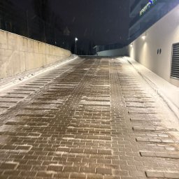 WE-GAM ALEKSANDER WĘGLARZ - Zimowy podjazd z kostki brukowej, lekko ośnieżony, prowadzący w górę w kierunku budynku z dekoracjami świątecznymi. Widoczne płatki śniegu i oświetlenie.