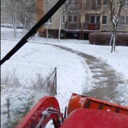 WE-GAM ALEKSANDER WĘGLARZ - Czerwony traktor odśnieża chodnik w zimowy dzień, widok z kabiny, budynki w tle.