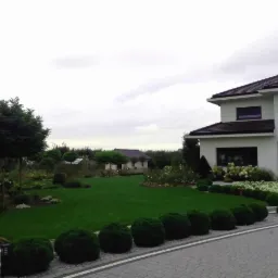 Zadbany ogród przydomowy z równo przystrzyżonym trawnikiem, kulistymi krzewami wzdłuż ścieżki i kwitnącymi rabatami, na tle nowoczesnego domu z ciemną dachówką w pochmurny dzień.
