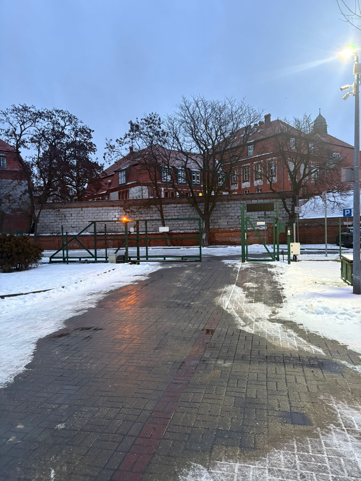 Zimowy widok na zieloną bramę wjazdową z ceglanym murem i budynkami w tle, oświetloną lampą. Śnieg leży na chodniku i trawniku.