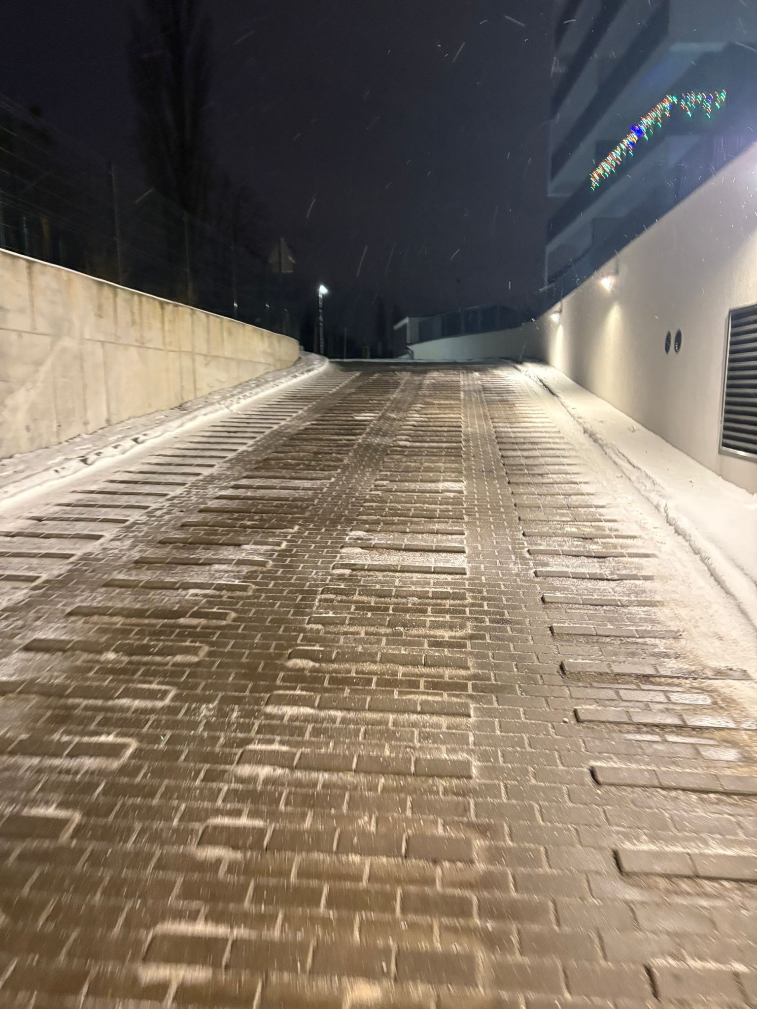 Zimowy podjazd z kostki brukowej, lekko ośnieżony, prowadzący w górę w kierunku budynku z dekoracjami świątecznymi. Widoczne płatki śniegu i oświetlenie.