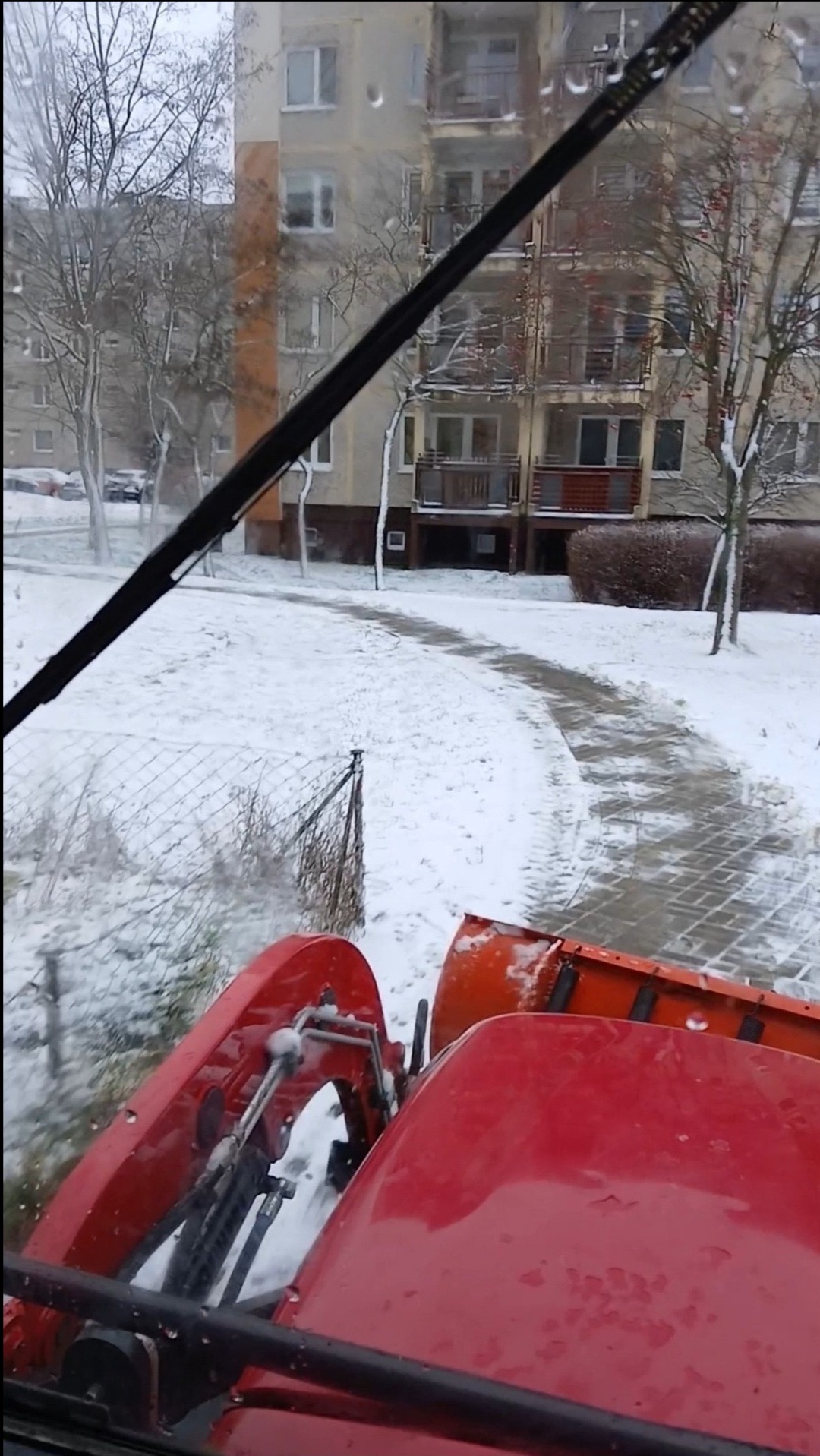 Czerwony traktor odśnieża chodnik w zimowy dzień, widok z kabiny, budynki w tle.