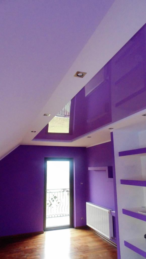 Liliowy pokój na poddaszu: ściany i sufit w odcieniach fioletu, białe elementy dekoracyjne. Widoczne okno balkonowe, grzejnik i regał z półkami.