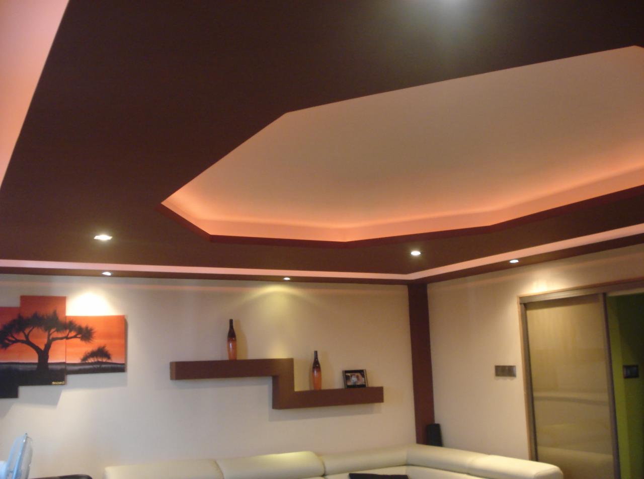 Nowoczesny salon z podwieszanym sufitem w odcieniach brązu i beżu, oświetlonym ledami. Dekoracyjne półki i obraz dodają wnętrzu charakteru.