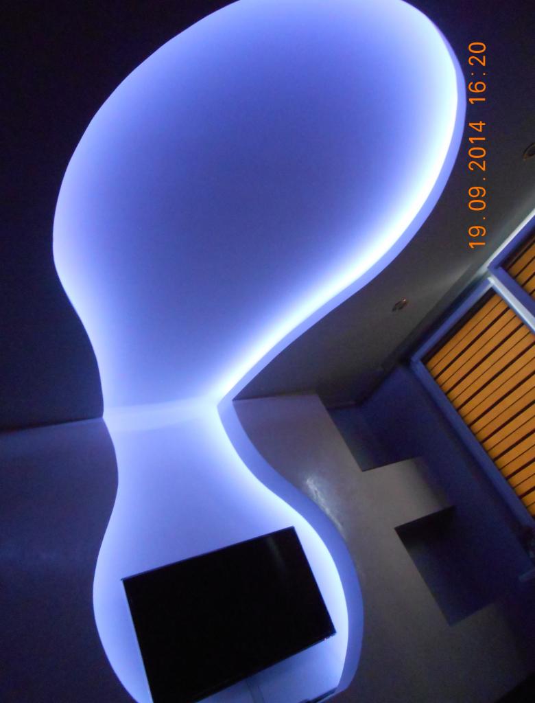 Nowoczesny, podwieszany sufit z oświetleniem LED w kształcie organicznym, integrujący się z ścianą i wnęką na telewizor. Efektowne, błękitne światło.