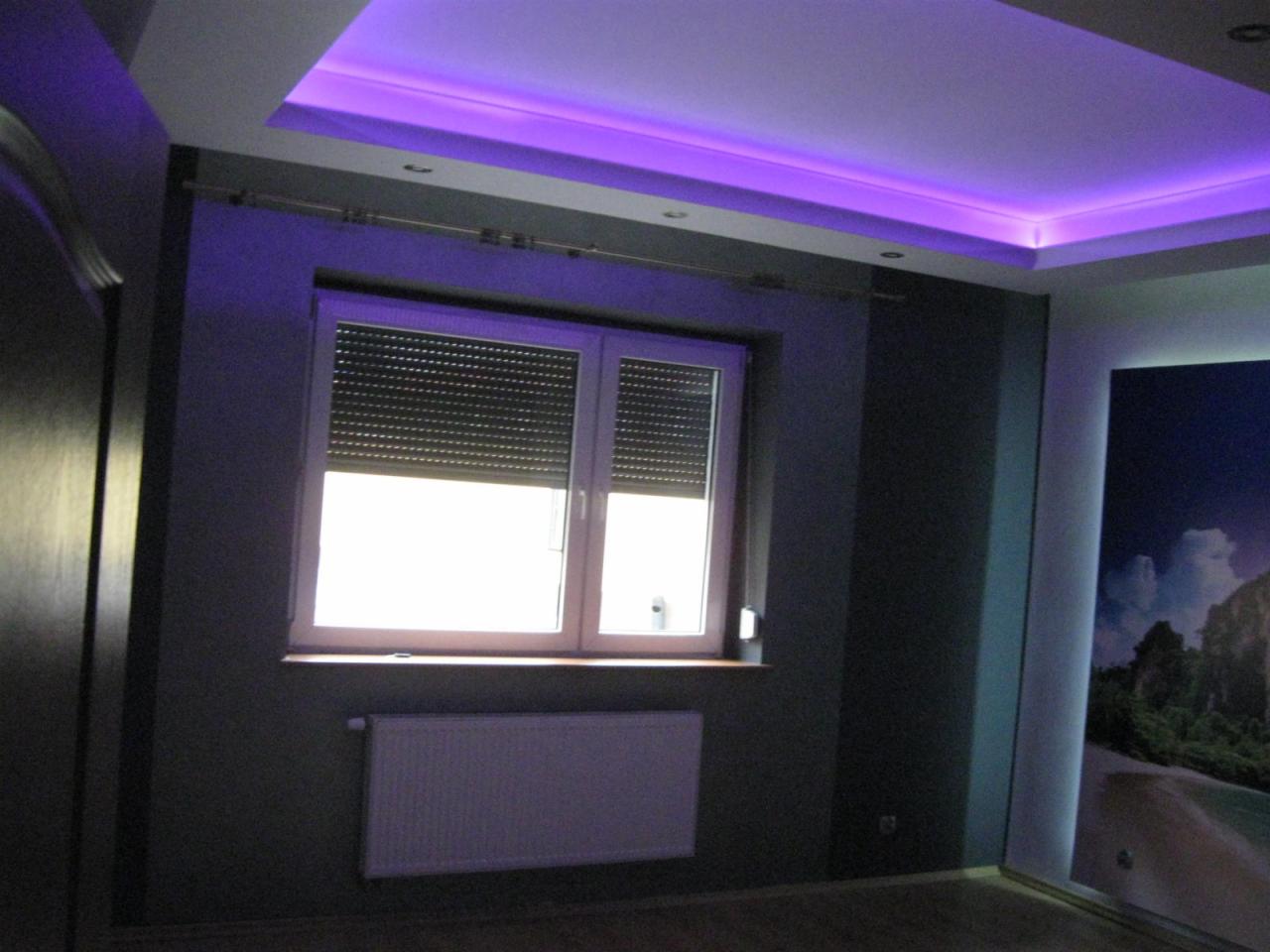 Pokój po remoncie: szare ściany, białe okno z roletami, grzejnik, podświetlany sufit na fioletowo, fototapeta z motywem przyrodniczym.