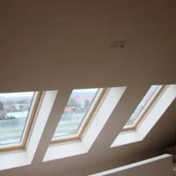 Trzy okna dachowe z drewnianymi ramami, ustawione pod skosem w jasnym pokoju na poddaszu. Za oknami widoczny krajobraz miejski w pochmurny dzień.