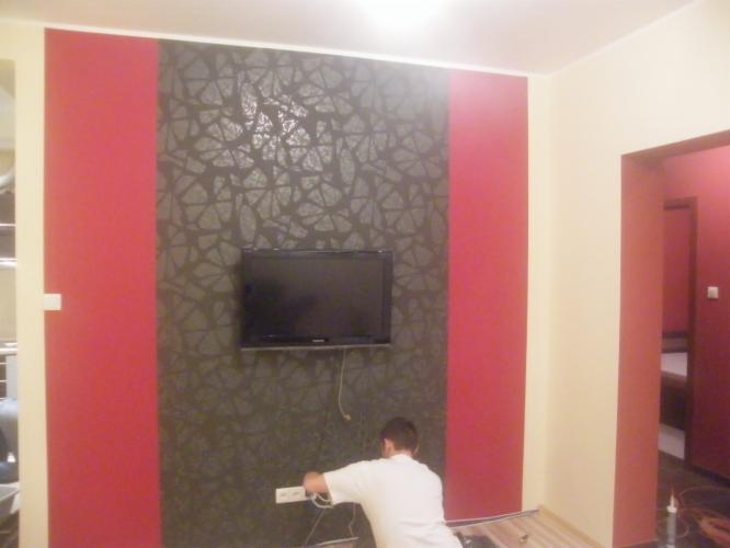 Wnętrze pokoju po remoncie: ściana z dekoracyjnym tynkiem w geometryczny wzór i czerwonymi pasami. Mężczyzna podłącza telewizor na ścianie, widoczne narzędzia.