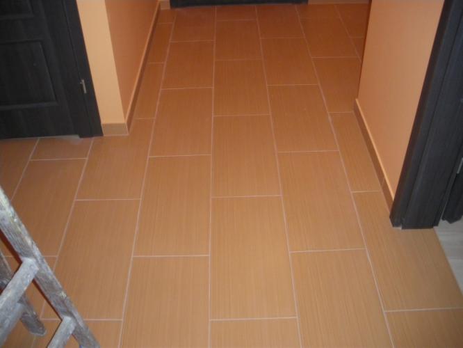 Ułożona, beżowa podłoga z płytek w korytarzu z ciemnymi drzwiami. Widoczny fragment drabiny. Jasne ściany. Ujęcie z góry, podkreślające wzór i precyzję ułożenia.