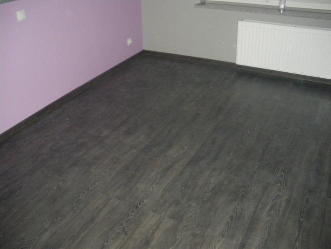 Ukończona podłoga z ciemnych paneli w pokoju z fioletową i szarą ścianą. Widoczny grzejnik pod oknem. Podłoga z wyraźną strukturą drewna.
