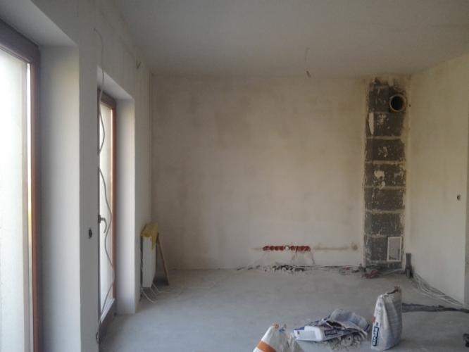 Surowy pokój po tynkowaniu: ściany pokryte tynkiem, widoczne instalacje elektryczne, worki z zaprawą na posadzce. Kolumna wentylacyjna z cegły.
