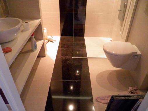 Wykończona łazienka z czarno-beżowymi płytkami podłogowymi i wiszącą toaletą. Widoczna umywalka z półkami i akcesoriami. Częściowo widoczne narzędzia na podłodze.