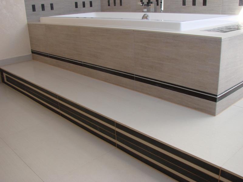 Estetyczne wykończenie łazienki z wanną obudowaną płytkami imitującymi drewno i dekoracyjnymi listwami. Podest wyłożony jasnymi płytkami z ciemnymi paskami.