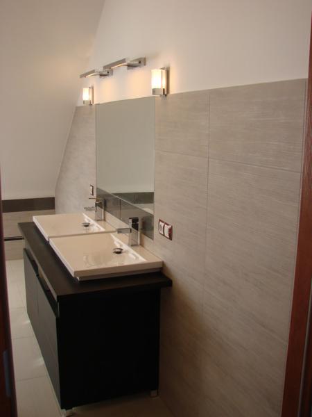 Nowoczesna łazienka z podwójną umywalką, minimalistycznym oświetleniem i płytkami imitującymi drewno na ścianie, widziana z perspektywy wejścia.