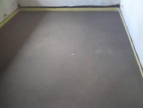 Gładka, szara posadzka jastrychowa w rogu pomieszczenia, zabezpieczona żółtą taśmą przy ścianach. Minimalistyczne wykończenie pod kolejne etapy prac.