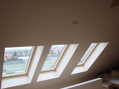 Trzy okna dachowe z drewnianymi ramami, ustawione pod skosem w jasnym pokoju na poddaszu. Za oknami widoczny krajobraz miejski w pochmurny dzień.