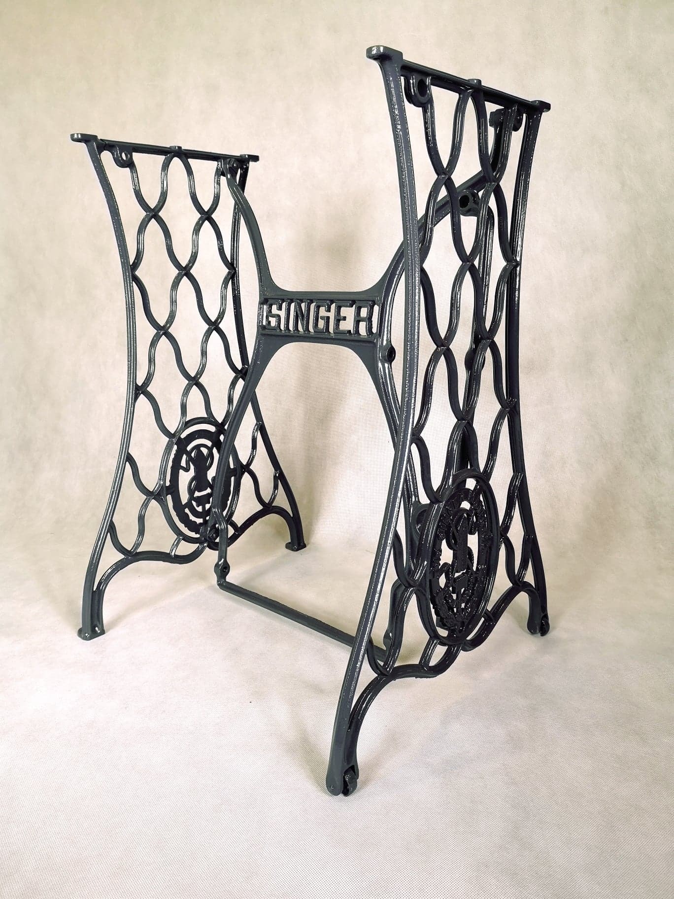 Odrestaurowana, żeliwna podstawa maszyny do szycia Singer z dekoracyjnymi elementami, świeżo pomalowana na ciemny kolor, eksponowana na jasnym tle.