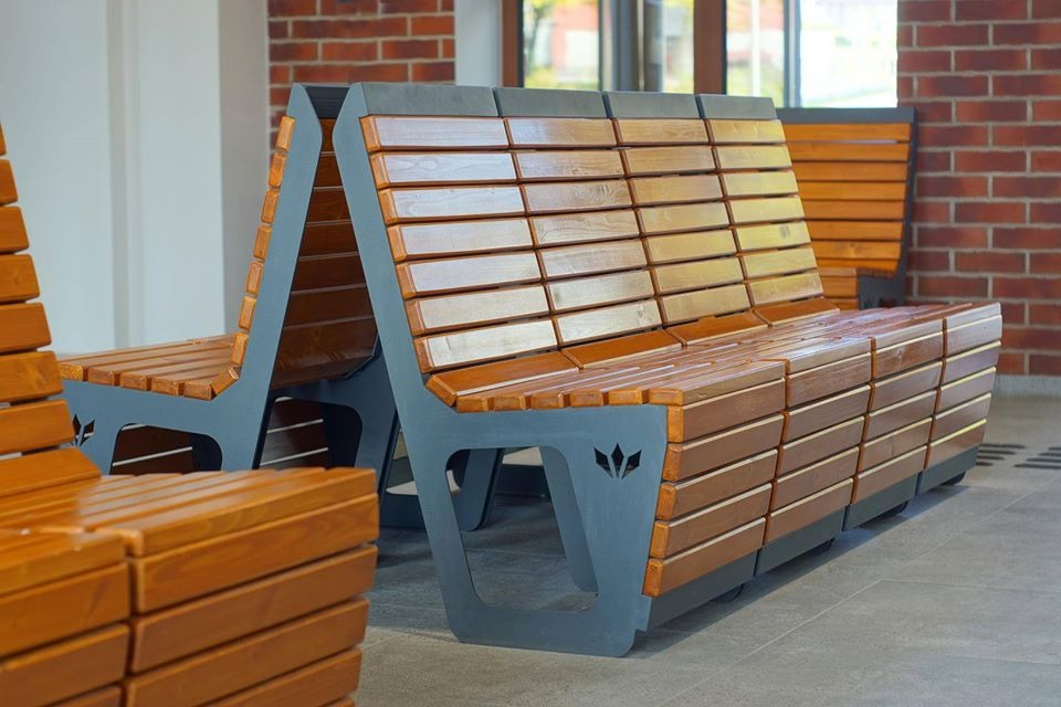 Nowoczesne, drewniane ławki parkowe z szarą, metalową konstrukcją, zdobione motywem roślinnym, ustawione na szarym chodniku przed budynkiem z cegły.