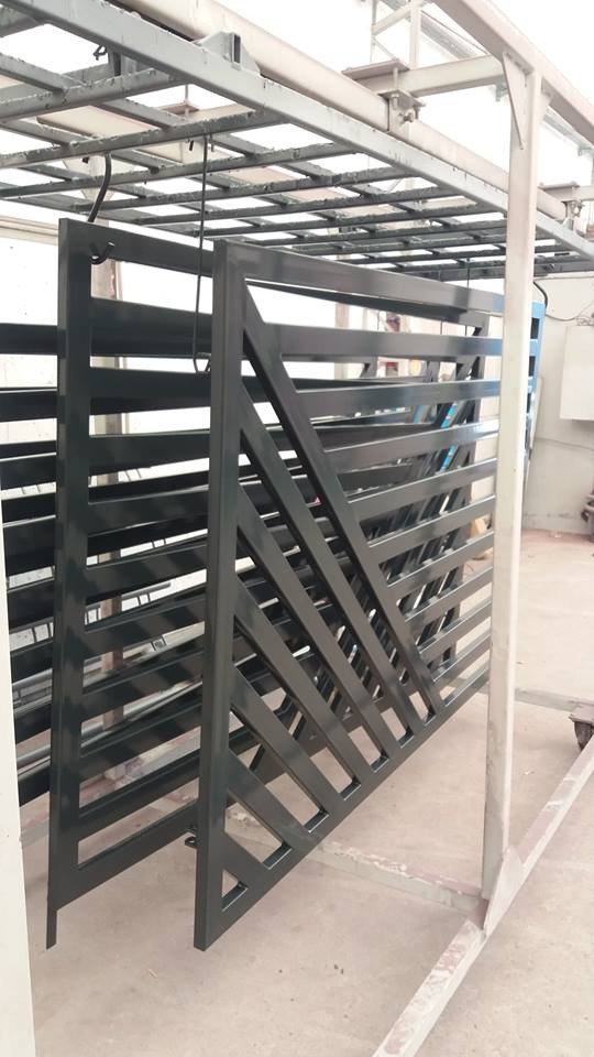 Metalowa brama ogrodzeniowa z poprzecznymi szczeblami, świeżo po nałożeniu farby, zawieszona na konstrukcji w hali produkcyjnej.