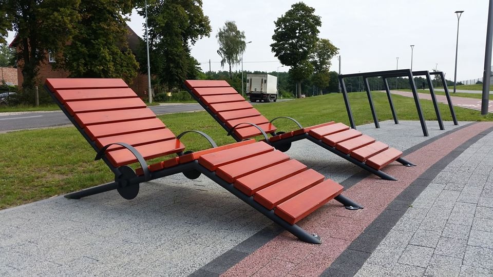 Dwa czerwone, drewniane leżaki miejskie z metalową konstrukcją na wybrukowanym chodniku, w tle zieleń i przejeżdżająca ciężarówka.