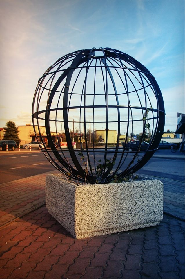 Metalowa konstrukcja w kształcie kuli, umieszczona w betonowej donicy na chodniku, z widoczną roślinnością wewnątrz, na tle ulicy i budynków.