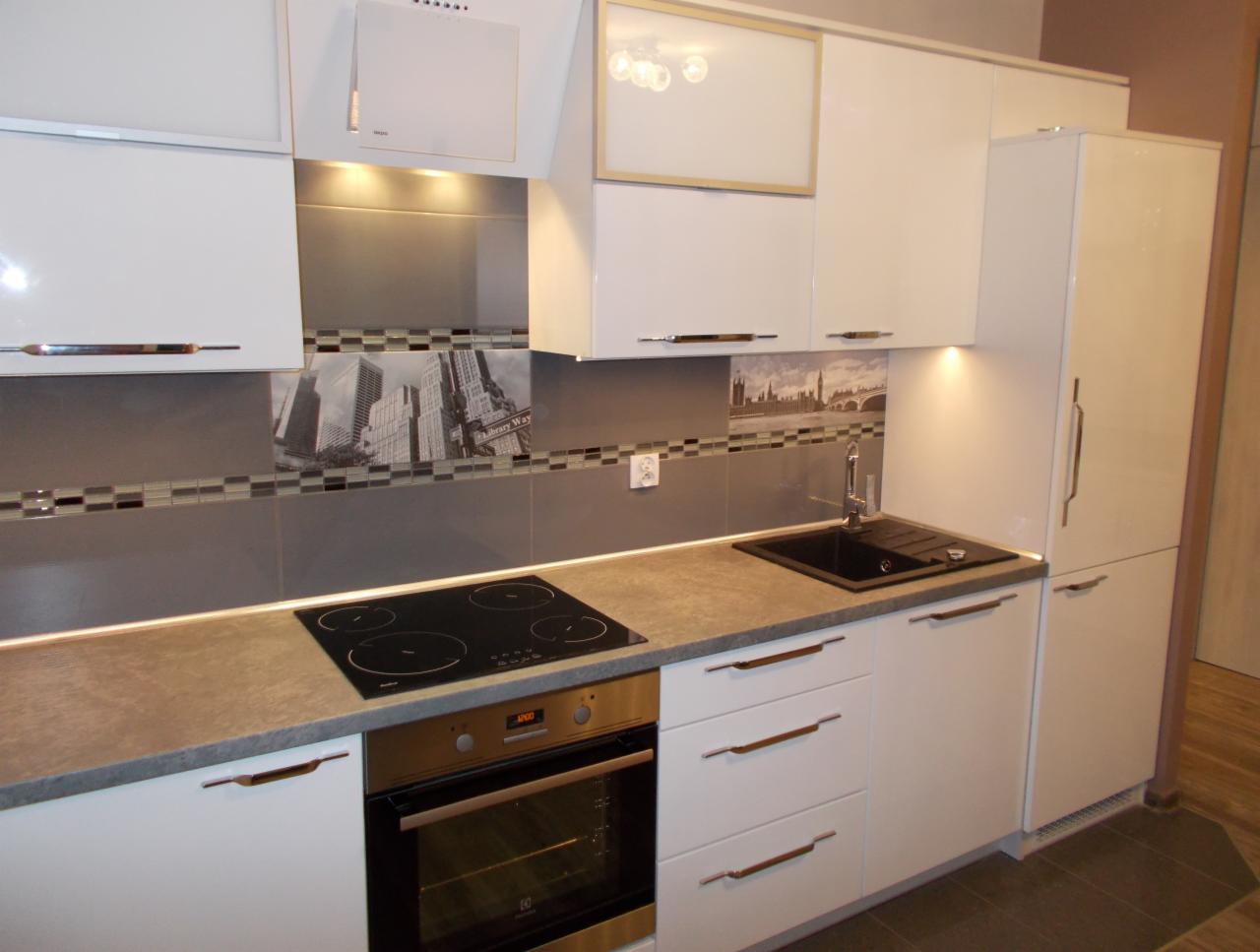 Biała zabudowa kuchenna z szarym blatem, płyta indukcyjna i piekarnik. Nad blatem dekoracyjne płytki z motywem miejskim i oświetlenie LED.