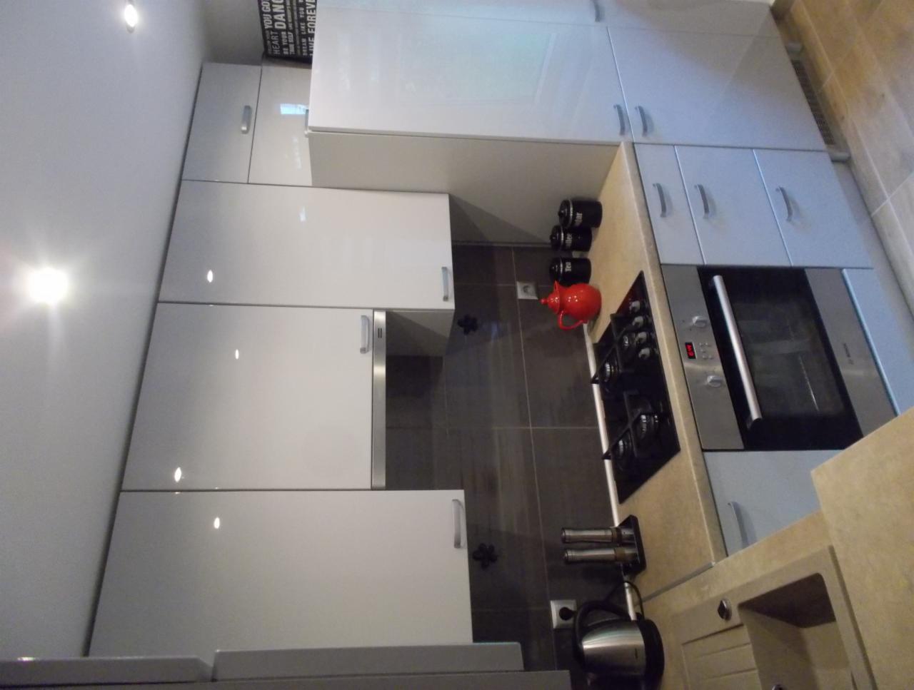 Biała, błyszcząca zabudowa kuchenna z piekarnikiem i płytą grzewczą. Na blacie stoją czarne pojemniki i czerwony czajnik. Widok na fragment ściany i sufit.