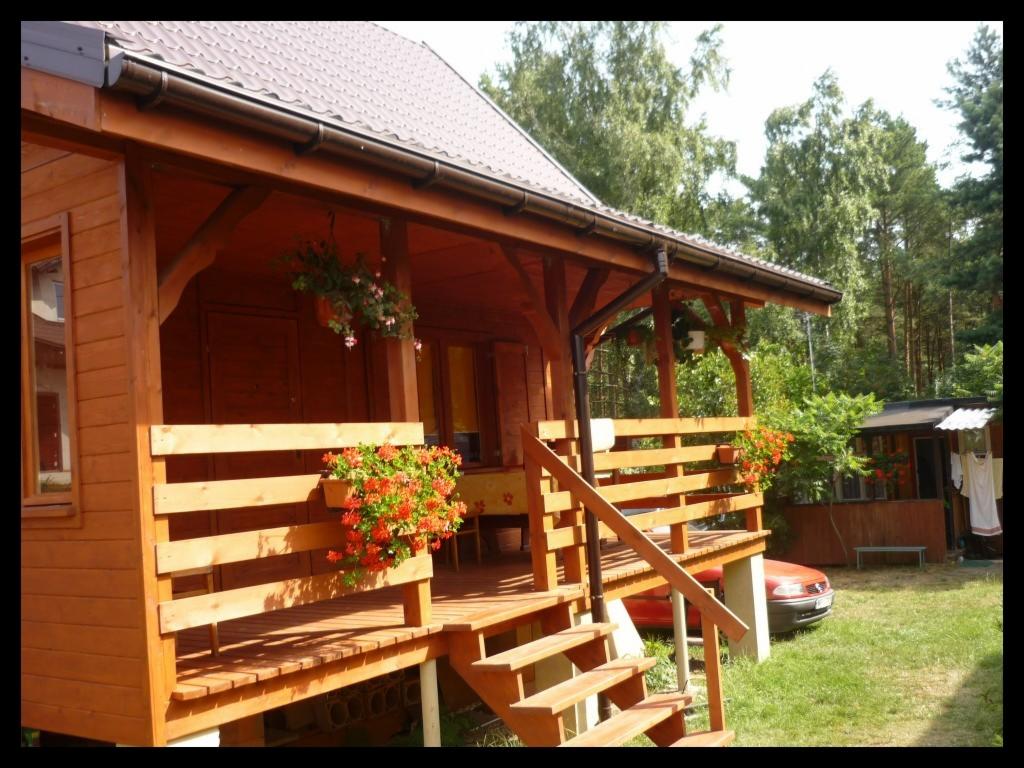 Drewniany domek letniskowy z werandą ozdobioną kwiatami w doniczkach i wiszących, brązowy dach z rynną, na trawniku częściowo widoczny czerwony samochód osobowy.