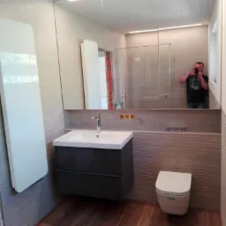 Wykończona łazienka z umywalką na szafce, podwieszaną toaletą i dużym lustrem z oświetleniem LED, widoczne także brązowe płytki podłogowe imitujące drewno oraz grzejnik naścienny. W lustrze odbija...