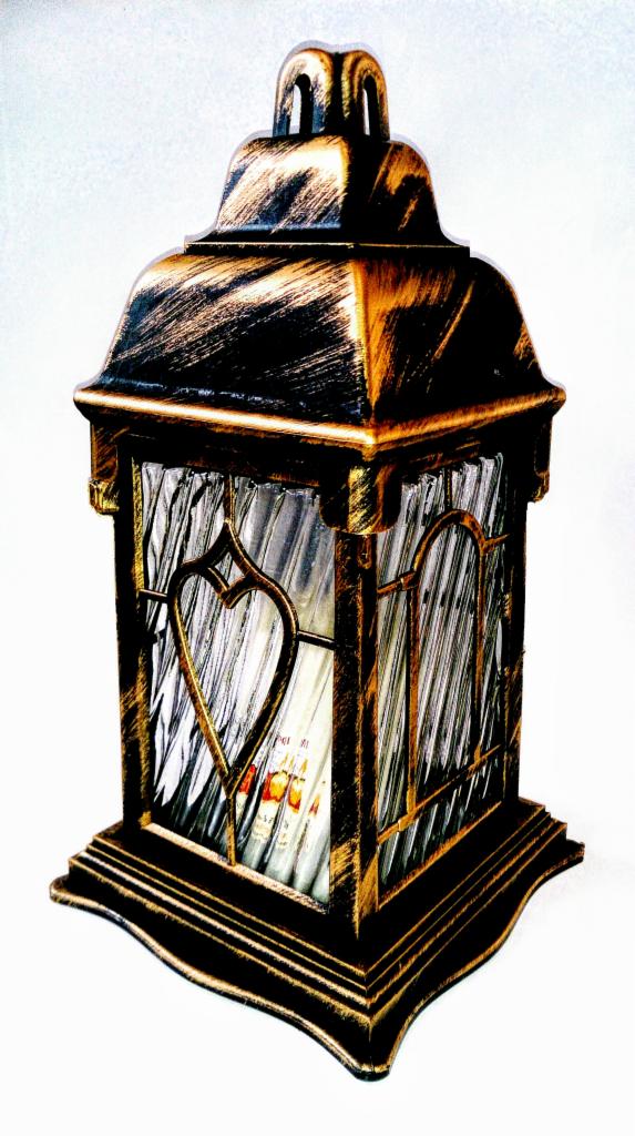 Ozdobny, metalowy lampion w kolorze starego złota z motywem serca na szklanych ściankach,  na białym tle. Dekoracja nagrobna.