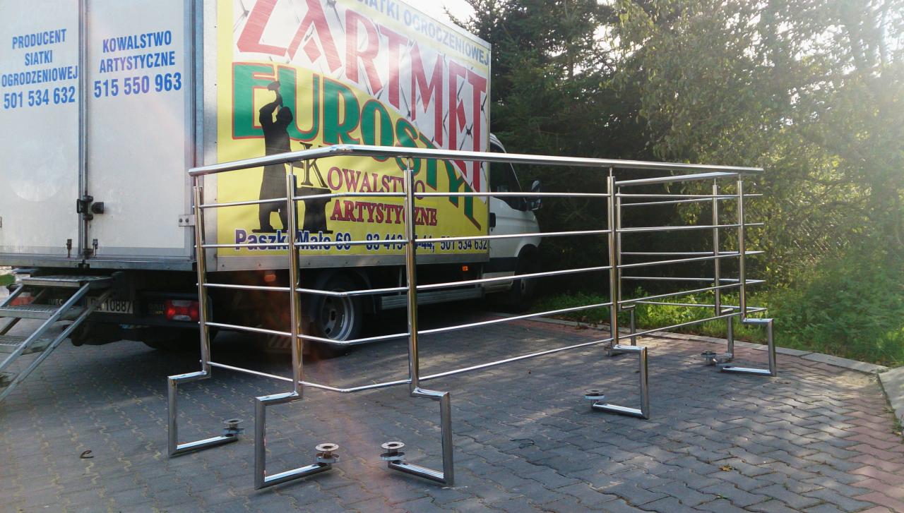 Konstrukcja z polerowanej stali nierdzewnej, prawdopodobnie fragment balustrady, na tle kostki brukowej i samochodu dostawczego z reklamą firmy.